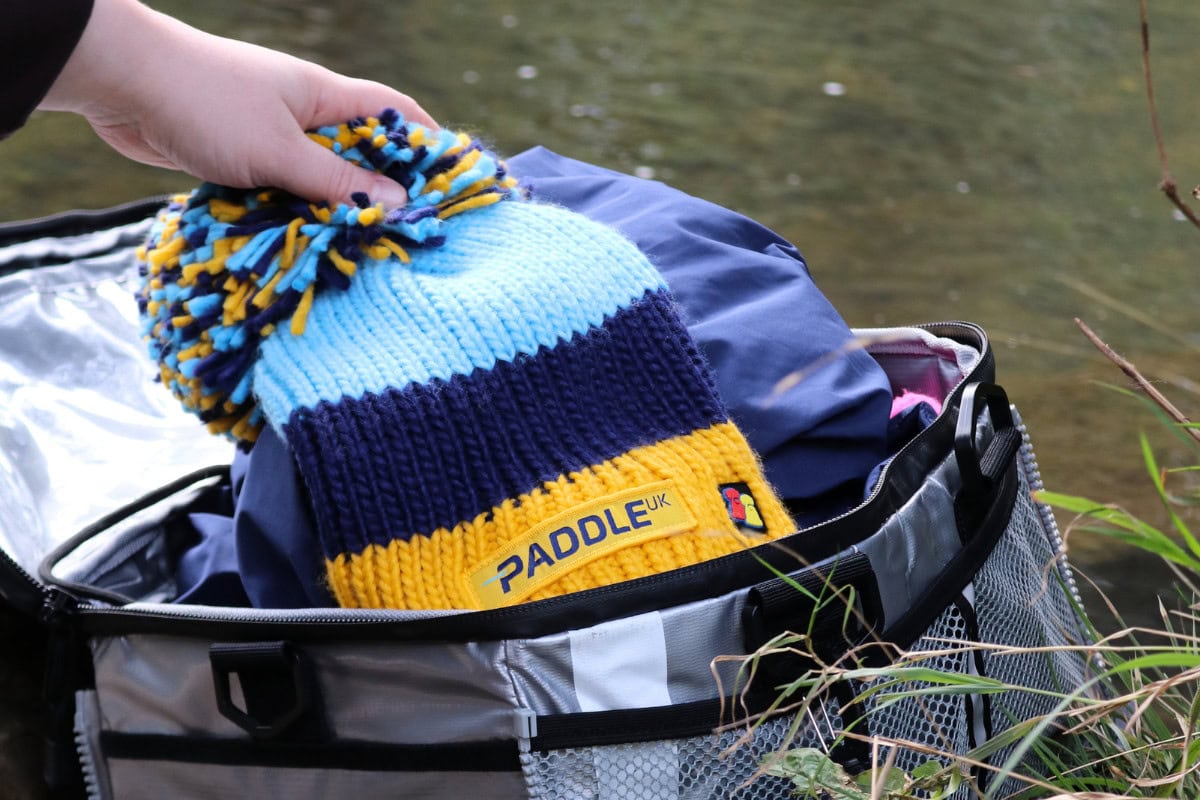 Paddle UK branded winter Big Bobble Hat being taken out of a KitBrix dry bag by the water. Essential kit to wear when paddling in autumn.