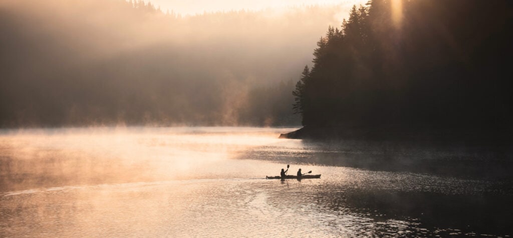 Two people in a two-person kayak paddling in autumn on a lake. Misty is coming off the water and the sun is setting through the trees.
