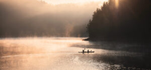 Two people in a two-person kayak paddling in autumn on a lake. Misty is coming off the water and the sun is setting through the trees.